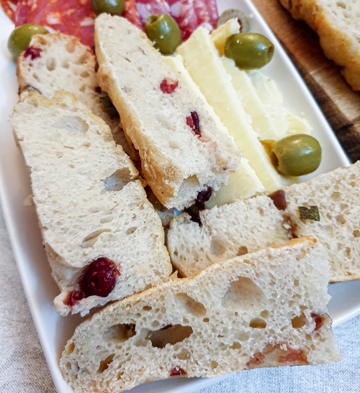 Slices of chestnut and cranberry focaccia on a white plate, with cheese, olives and slices salami.