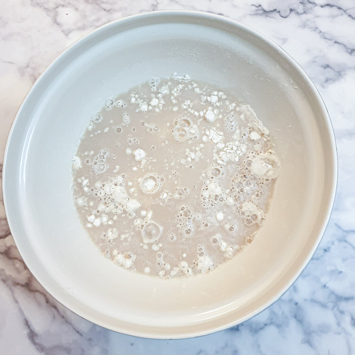 Yeast and water poured over flour in a large white mixing bowl.