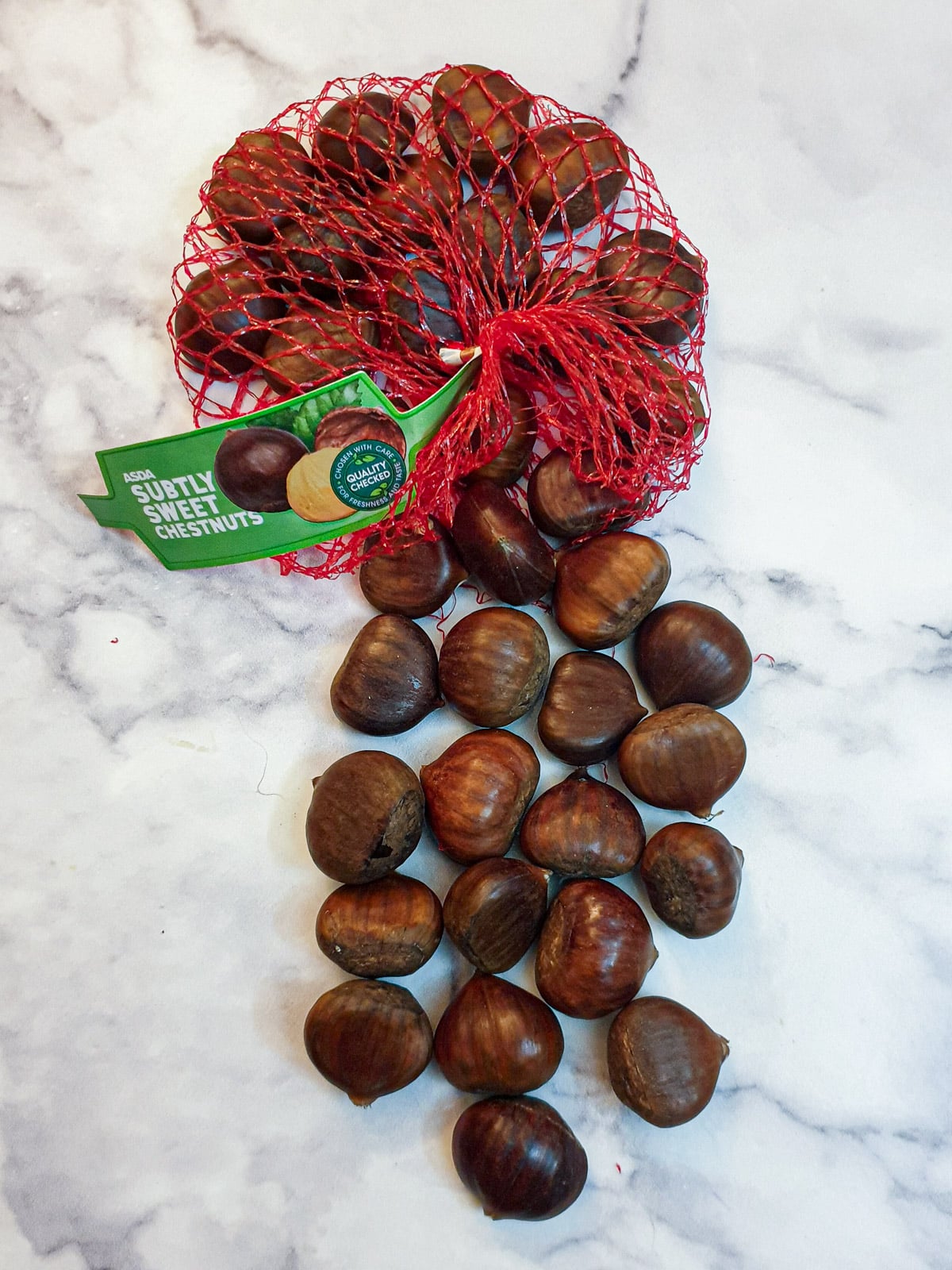 A red net bag of chestnuts, on a marble worktop with some of the chestnuts spilling out.
