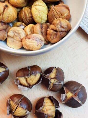 A dish of peeled roast chestnuts with 6 unpeeled roasted chestnuts in the foreground.