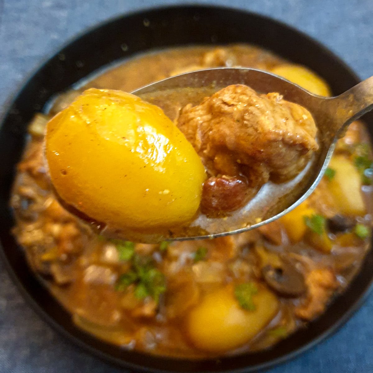 A potato and a piece of pork on a spoon held about a dish of pork casserole.
