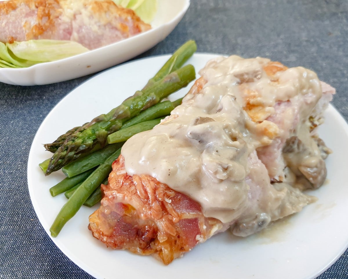 A bacon-wrapped chicken breast on a white plate, smothered in mushroom sauce with a side of asparagus spears.