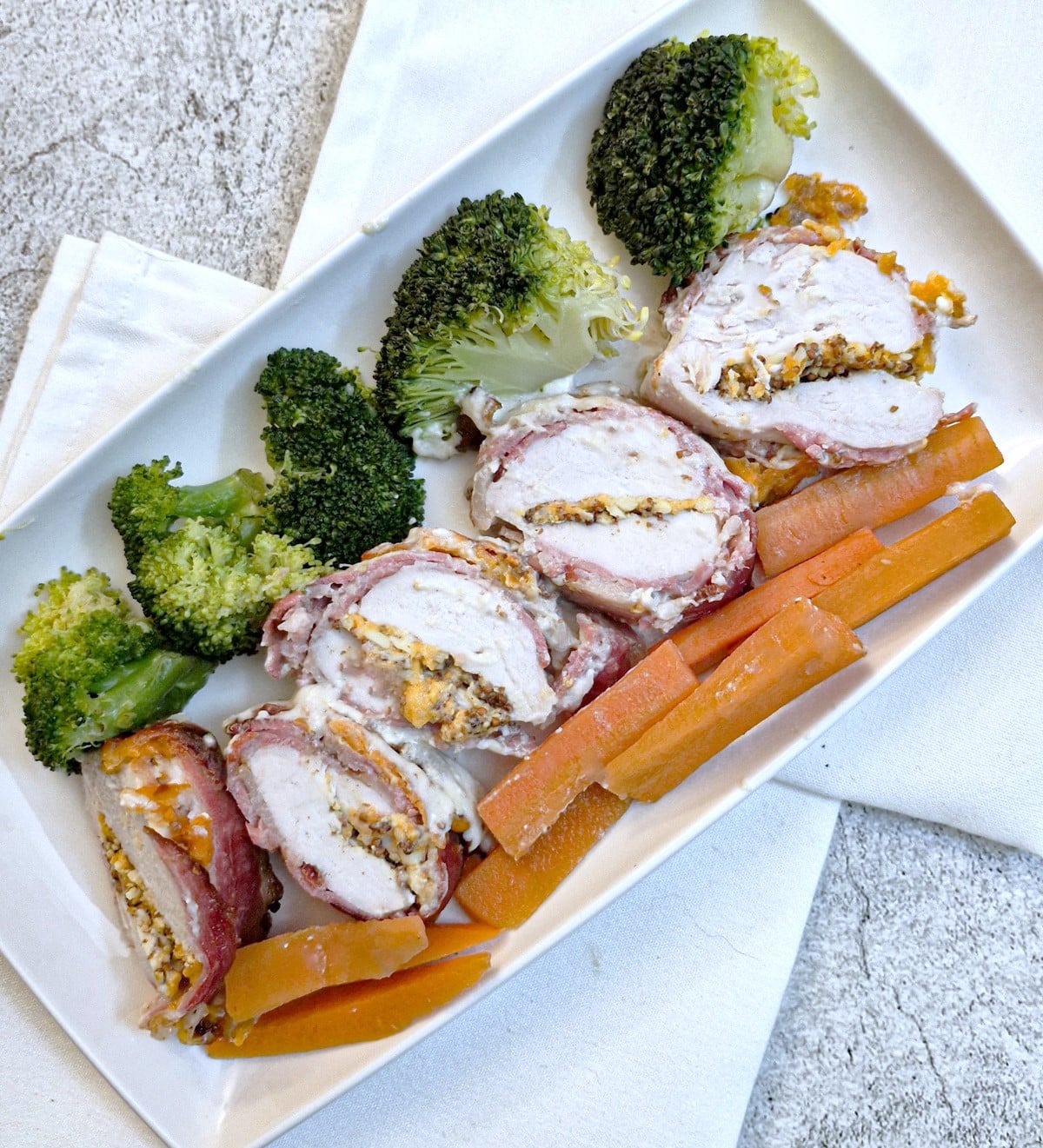 A white serving platter with slices of bacon wrapped chicken breast surrounded by broccoli and carrots.