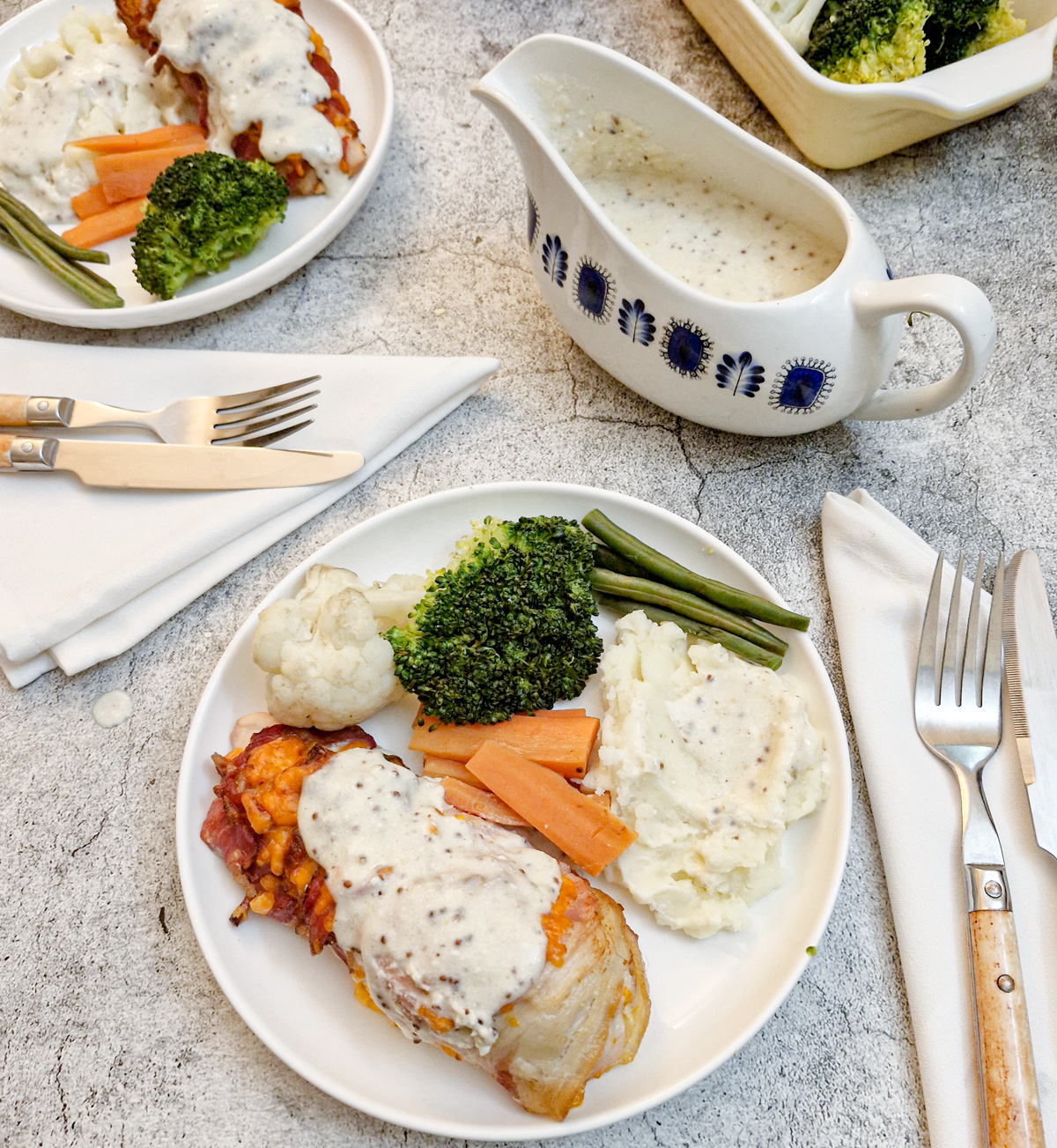 Overhead shot of a plate of bacon wrapped chicken breast with vegetables.  There is a blue patterned jug in the background containing  a creamy sauce.