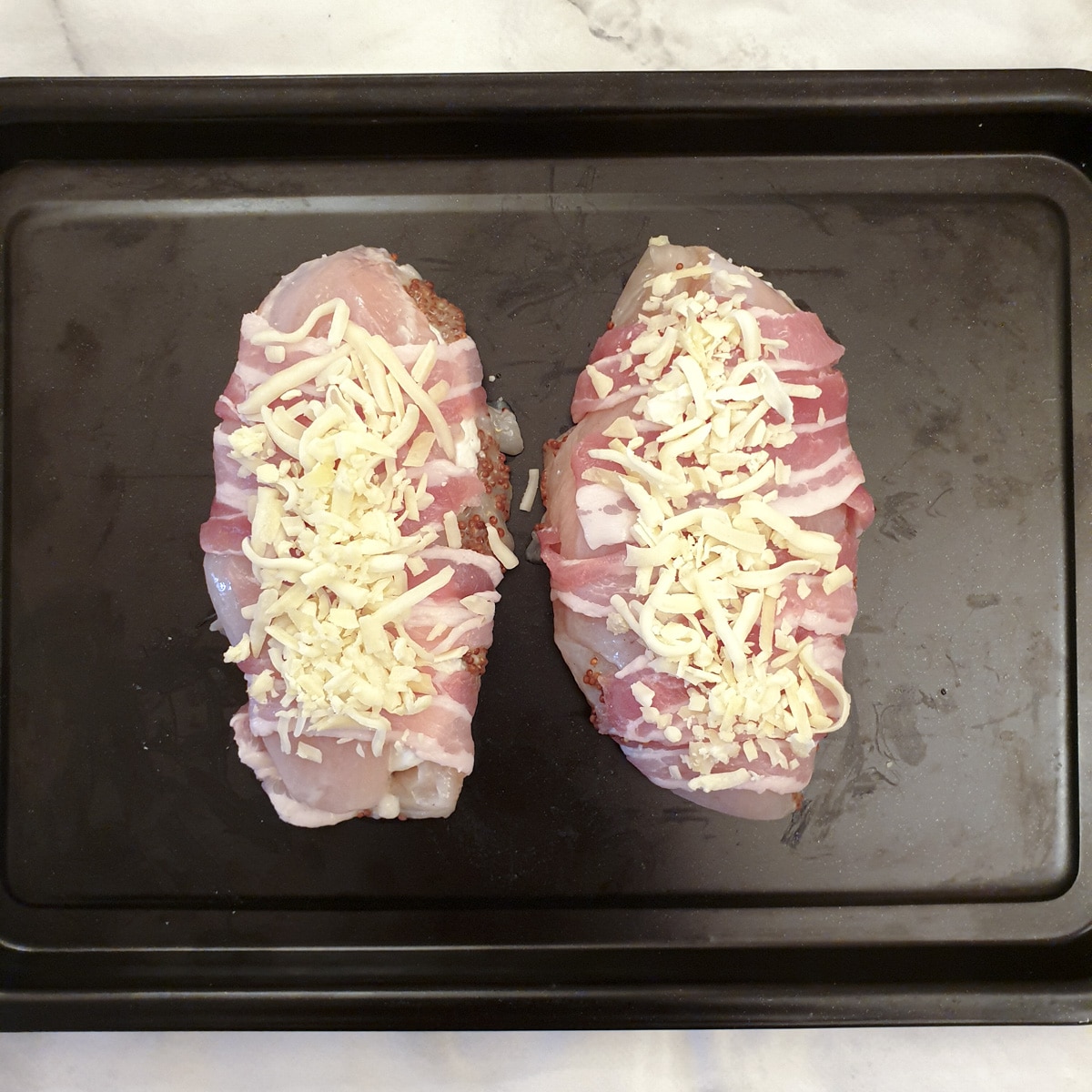 Two bacon wrapped chicken breasts sprinkled with grated cheese on a baking tray.