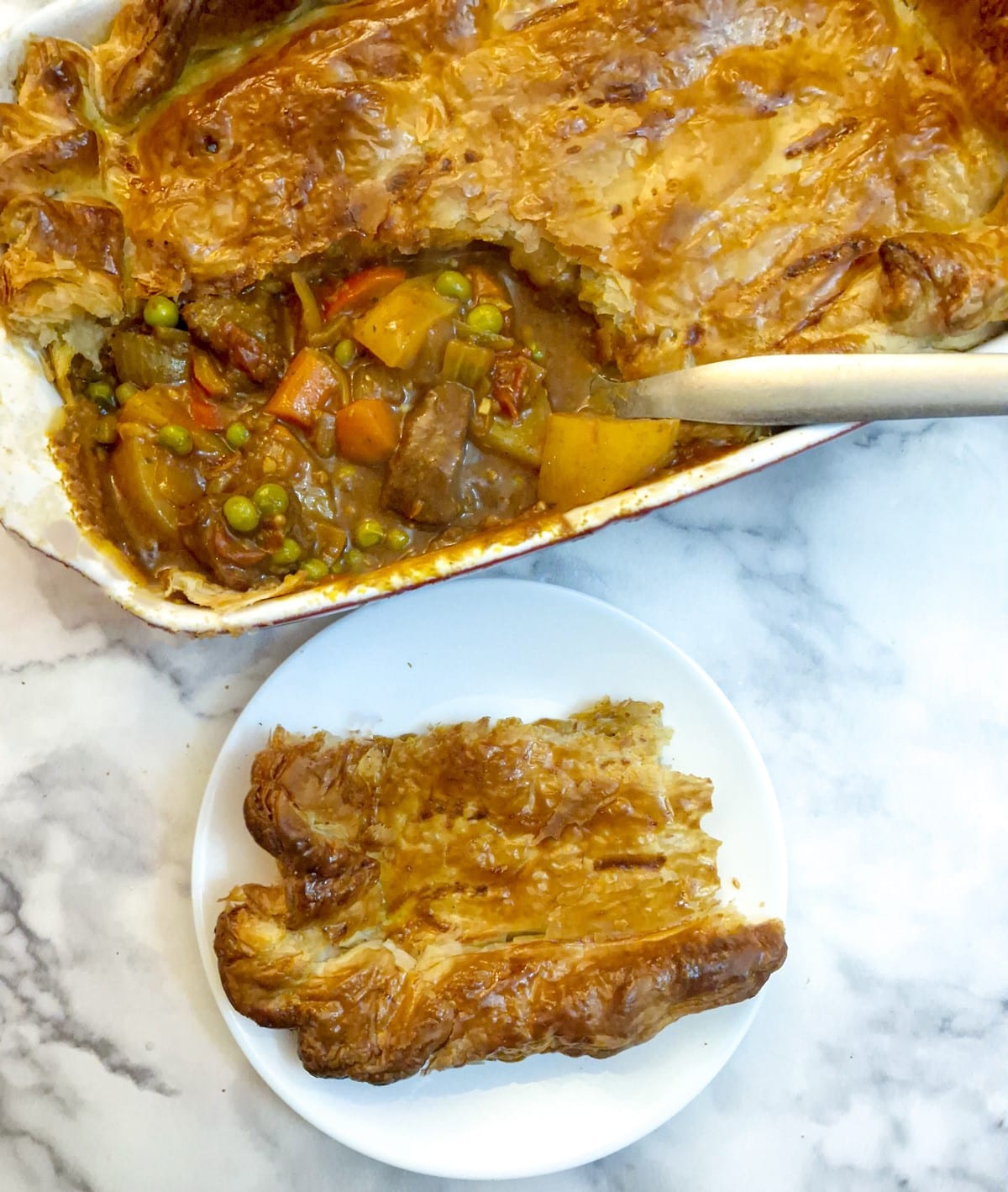 Overhead shot of beef curry pie with a corner of the pastry removed onto a side plate.  There is a spoon in the pie dish in the filling.