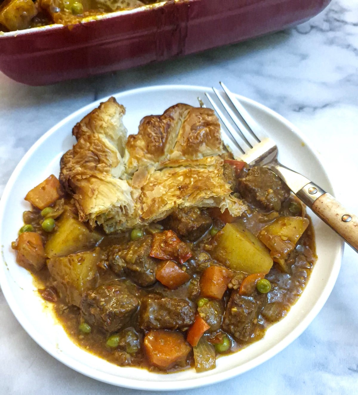 A helping of beef curry pie topped with crispy puff pastry on a white plate.