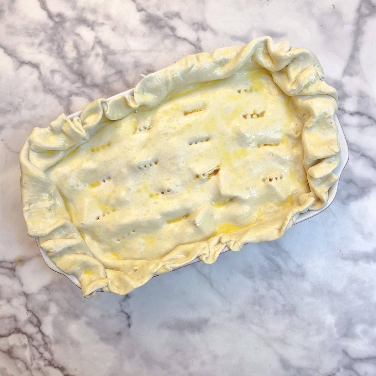 Overhead shot of the unbaked pie with the edges of pastry scrunched around the sides, and a coating of egg-wash on top.