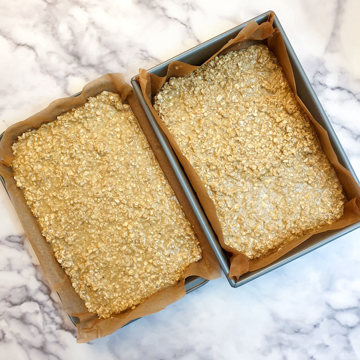Two baking trays lined with baking parchment and filled with unbaked flapjacks.