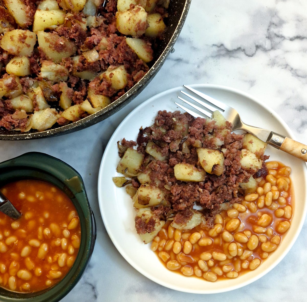 Corned beef hash on a white plate with a helping of baked beans. The pan of corned beef has and a dish of baked beans is also in the photo.