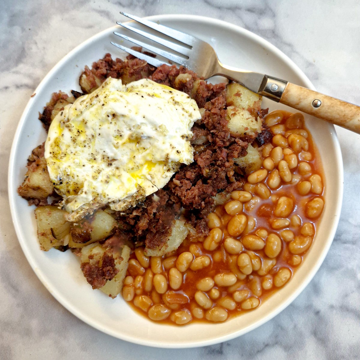 A white plate with a helping of corned beef hash and a side of baked beans, topped with a fried egg.