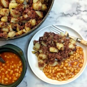 Corned beef hash on a white plate with a helping of baked beans. The pan of corned beef has and a dish of baked beans is also in the photo.