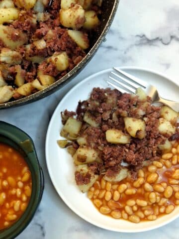 Corned beef hash on a white plate with a helping of baked beans. The pan of corned beef has and a dish of baked beans is also in the photo.