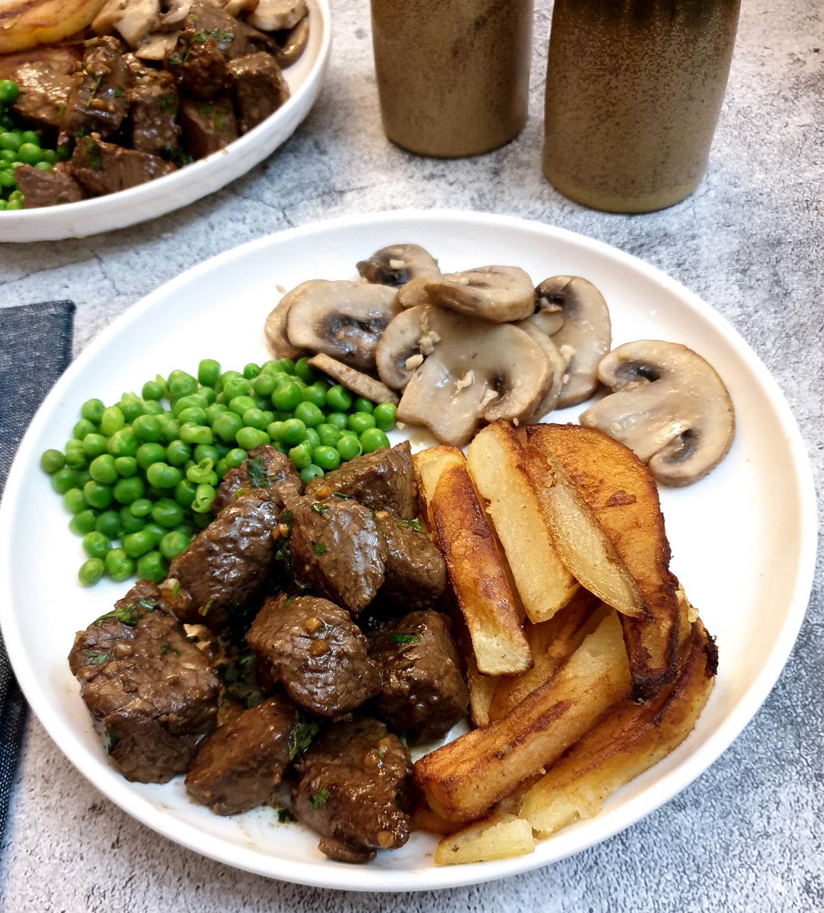 A white plate with garlic butter steak bites, chips, peas and garlic mushrooms.