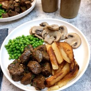 A white plate with garlic butter steak bites, chips, peas and garlic mushrooms.