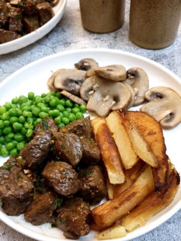 A white plate with garlic butter steak bites, chips, peas and garlic mushrooms.