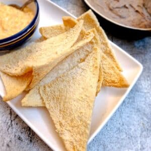 Slices of melba toast on a white plate with bowls on dips in the background.