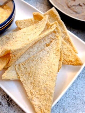 Slices of melba toast on a white plate with bowls on dips in the background.