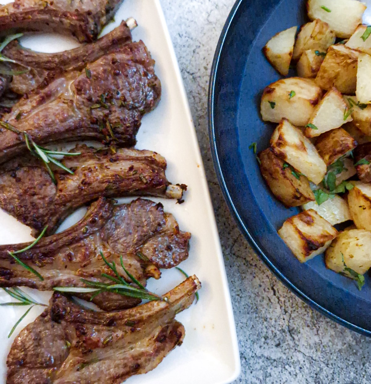 A white serving plate with 5 lamb chops, next to a dish of roast potatoes.