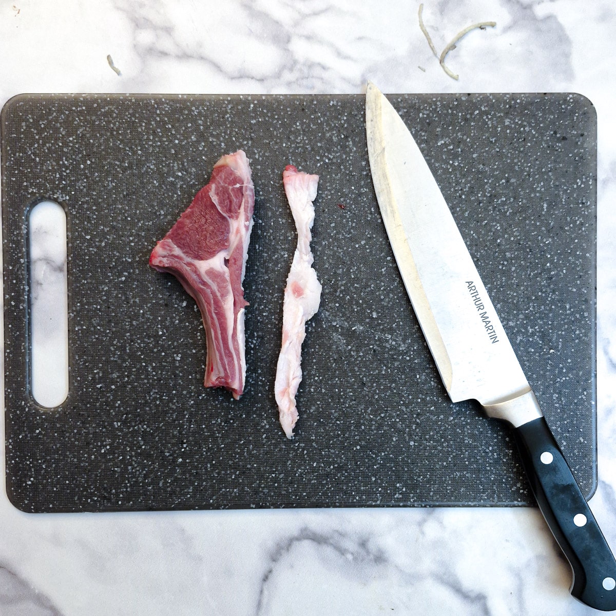 A lamb chop on a chopping board alongside a strip of fat which has been removed.  There is also a knife on the chopping board.