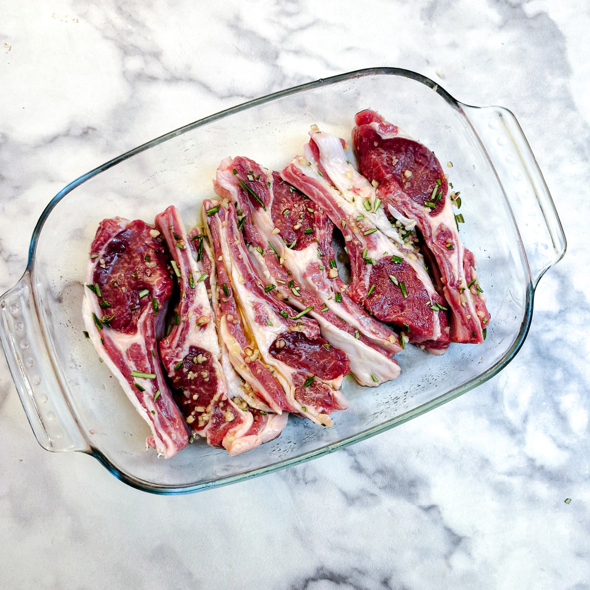 Six lamb rib chops covered in a marinade rub, in a shallow glass dish.