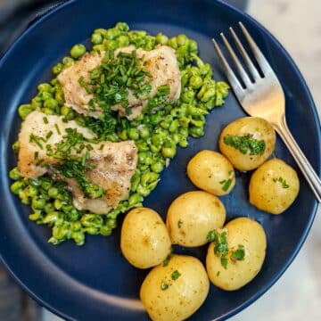 A blue plate with a helping of smashed minted peas topped with two pieces of monkfish, garnished with chives. There is also a small pile of baby potatoes on the plate.