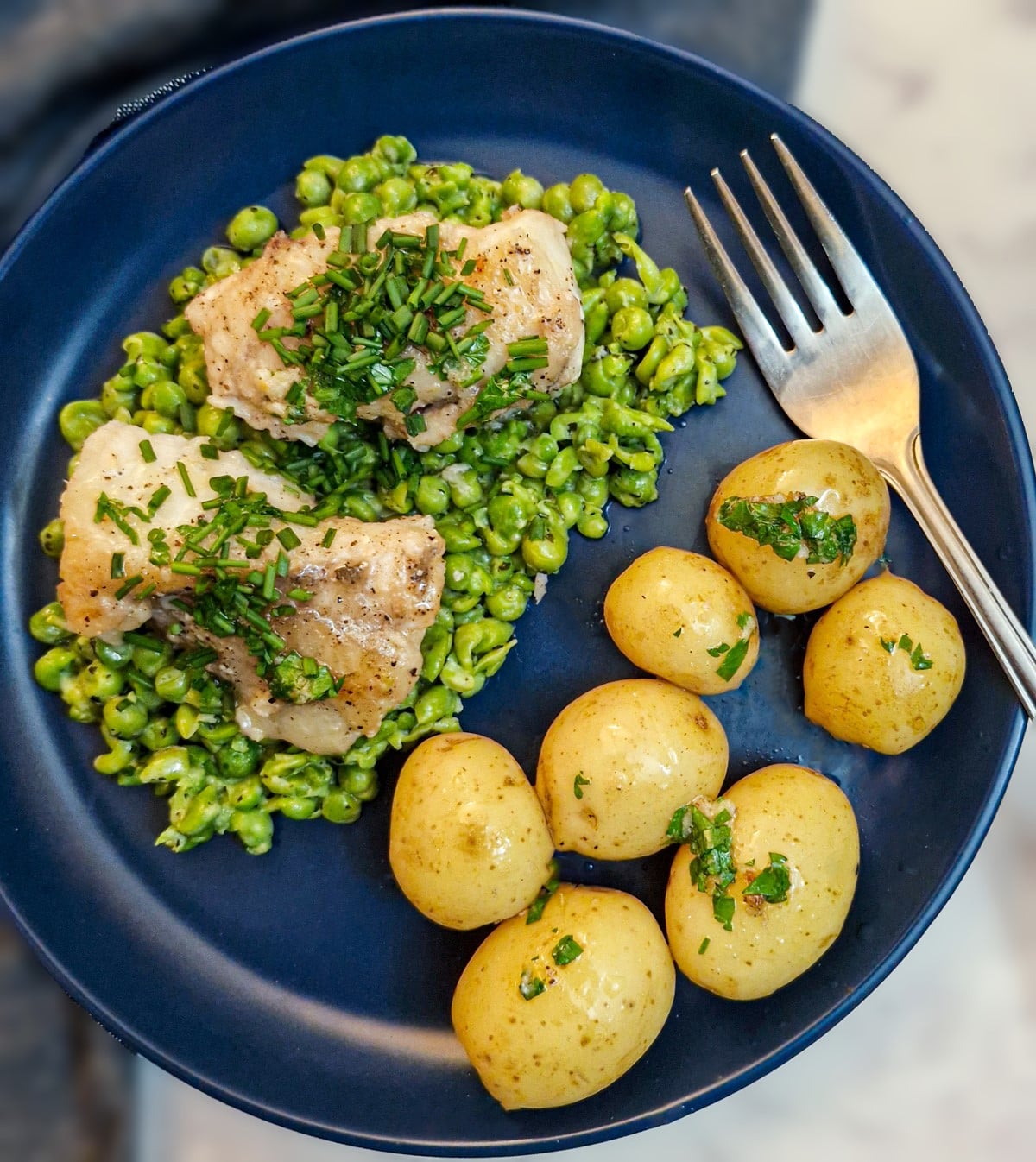 A blue plate with a helping of smashed minted peas topped with two pieces of monkfish, garnished with chives. There is also a small pile of baby potatoes on the plate.