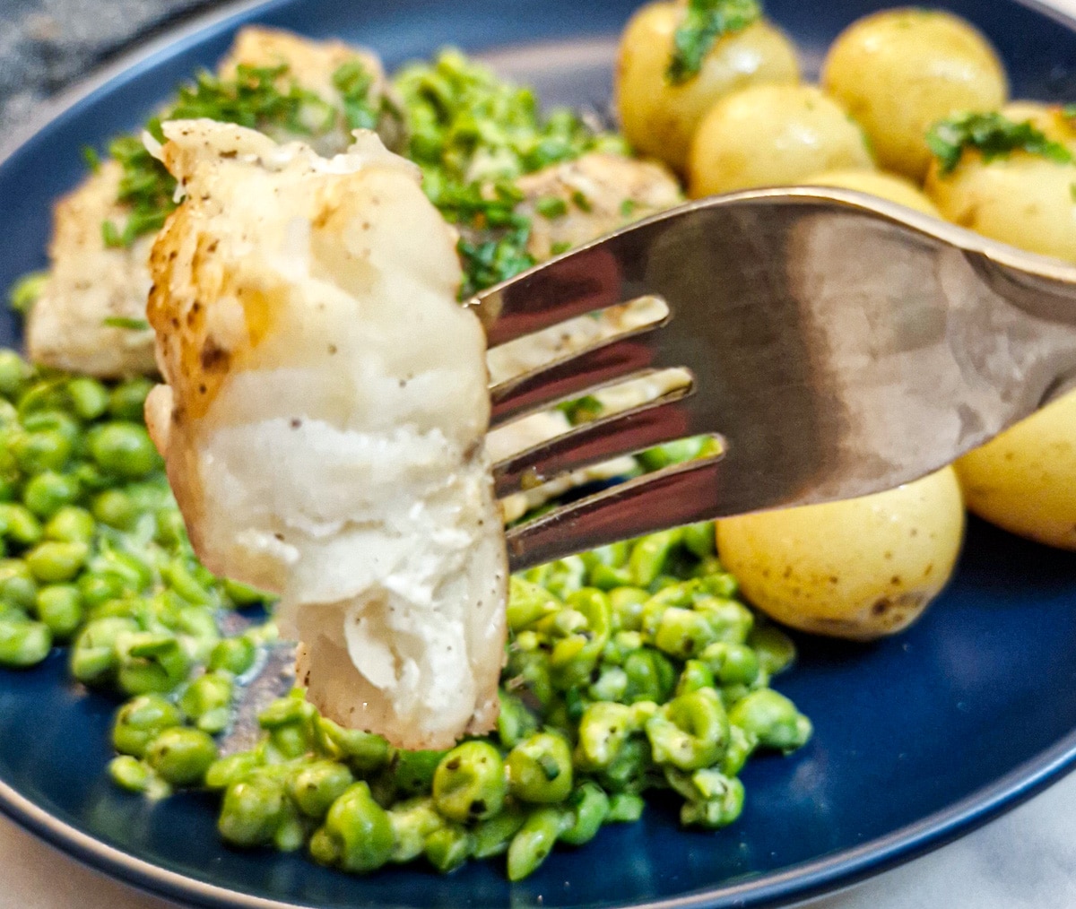 Close up of a piece of monkfish on a fork with a plate of peas and potatoes in the background.