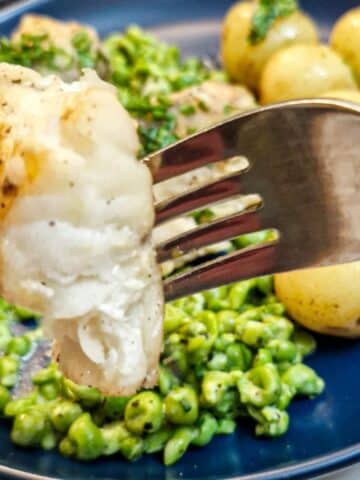 Close up of a piece of monkfish on a fork with a plate of peas and potatoes in the background.