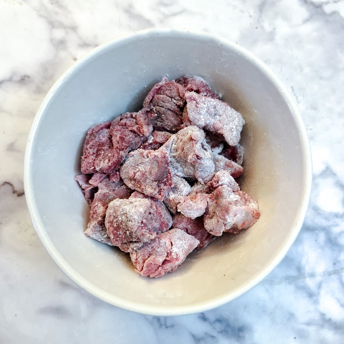 A small white bowl containing pieces of beef which have been coated in seasoned flour.