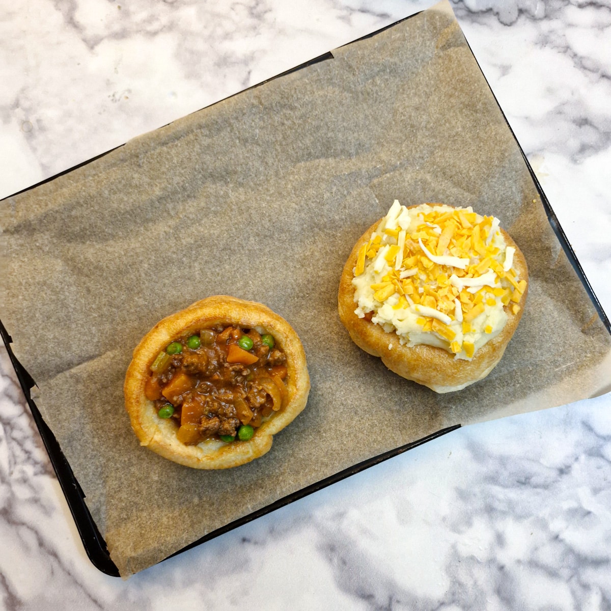 2 Yorkshire puddings on a baking tray, one stuffed with savoury mince and the other covered with mashed potatoes and grated cheese.