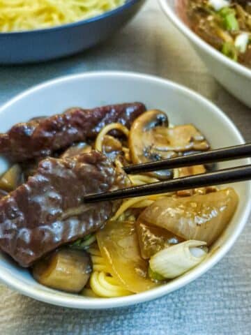 A piece of beef held with chopsticks over a bowl of beef and mushroom stirfry.