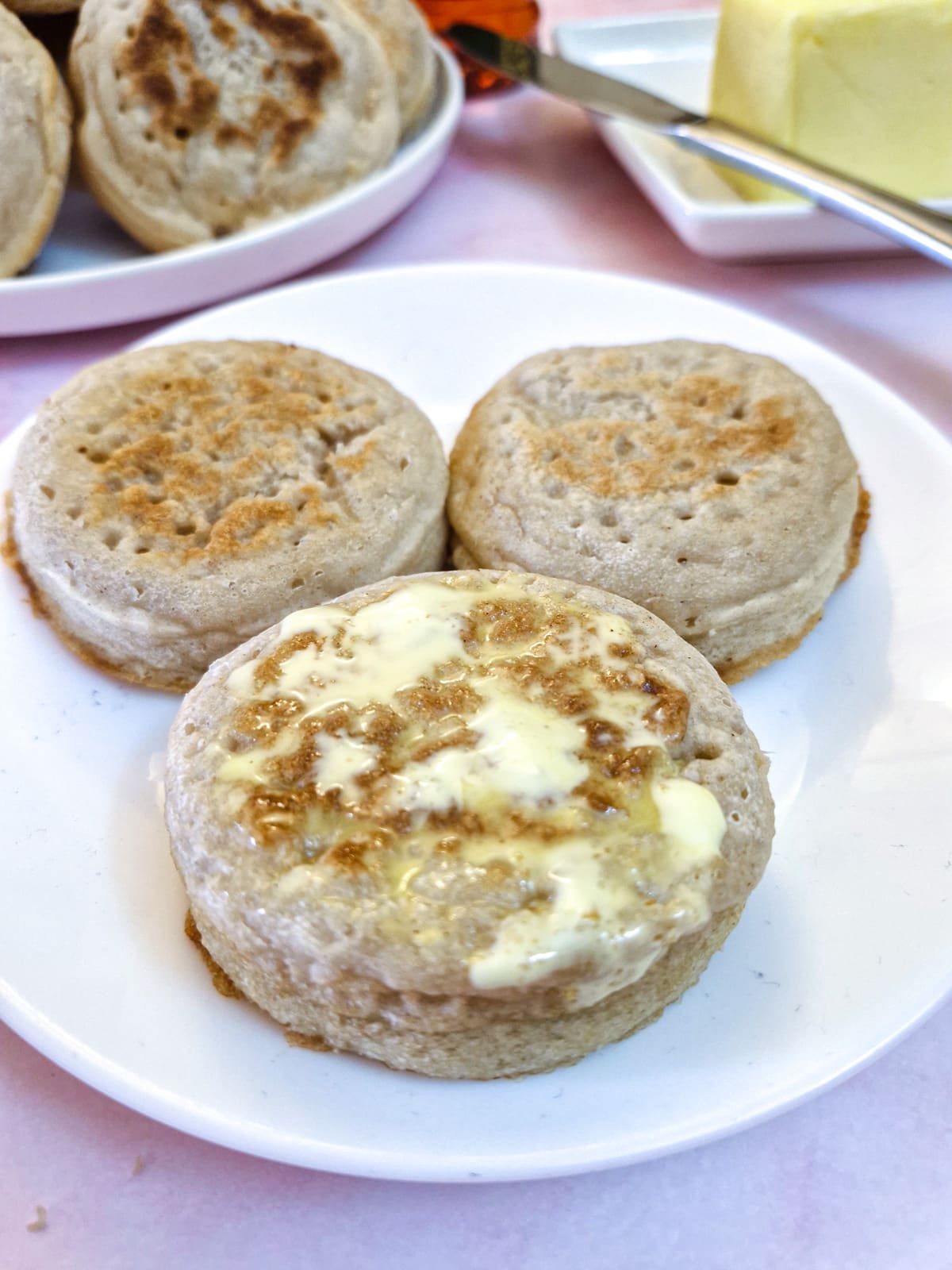 Three crumpets on a white plate, one of them spread with butter that is melting.