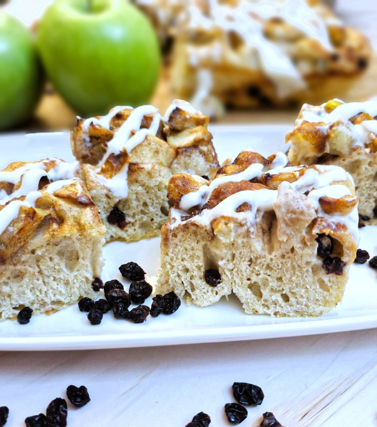 4 slices of apple and raisin focaccia on a white plate with apples in the background.