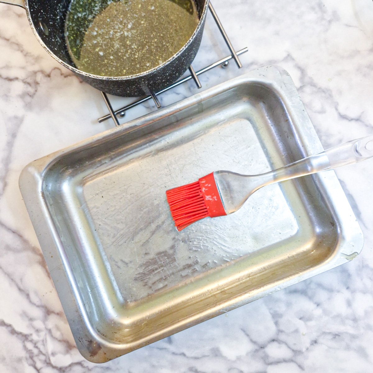 An oblong metal baking dish being brushed with melted butter.