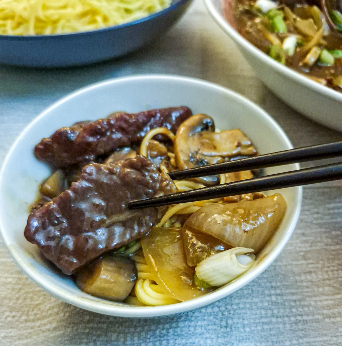 A piece of beef held with chopsticks over a bowl of beef and mushroom stirfry.