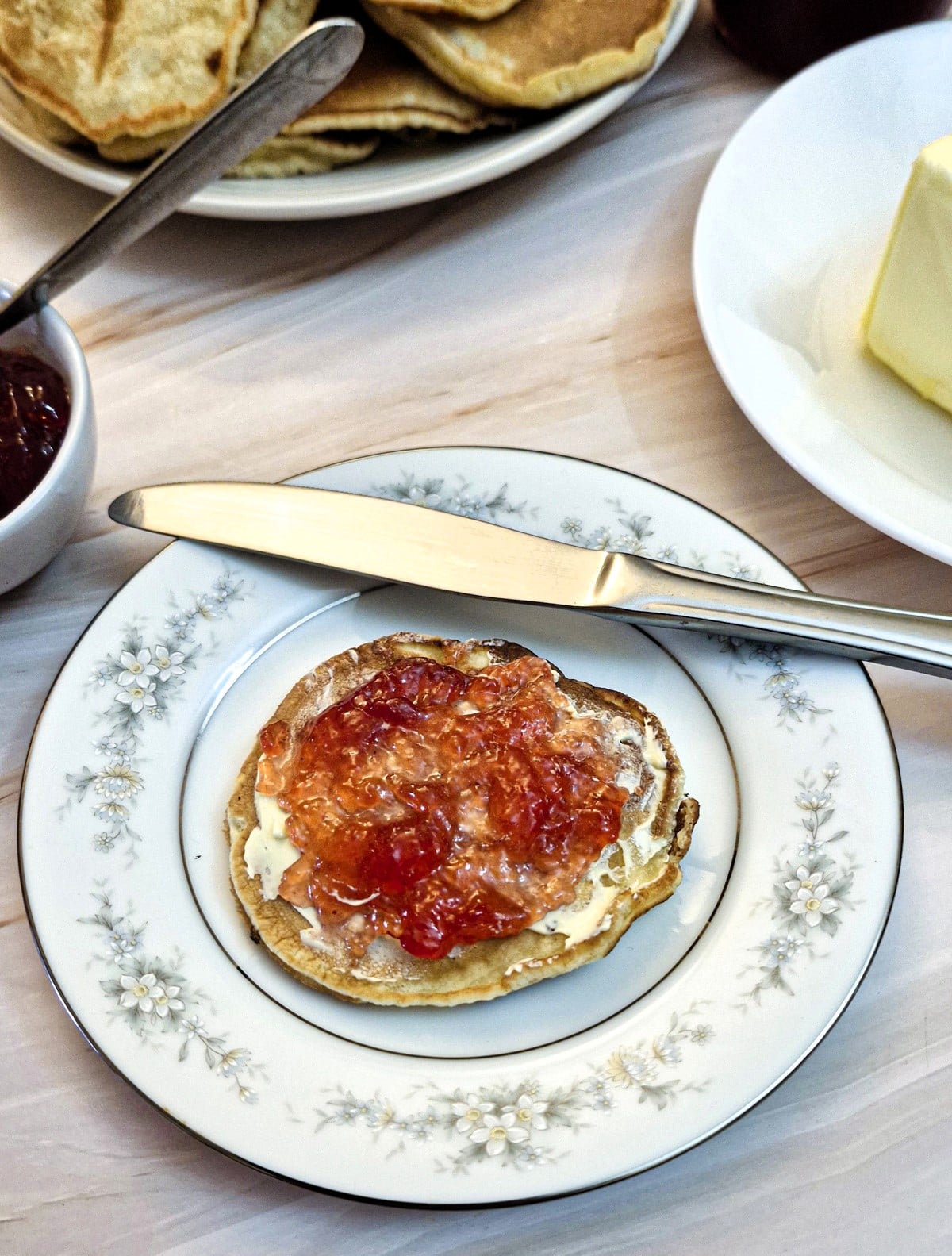 A drop scone spread with butter and strawberry jam on a white plate with a knife.