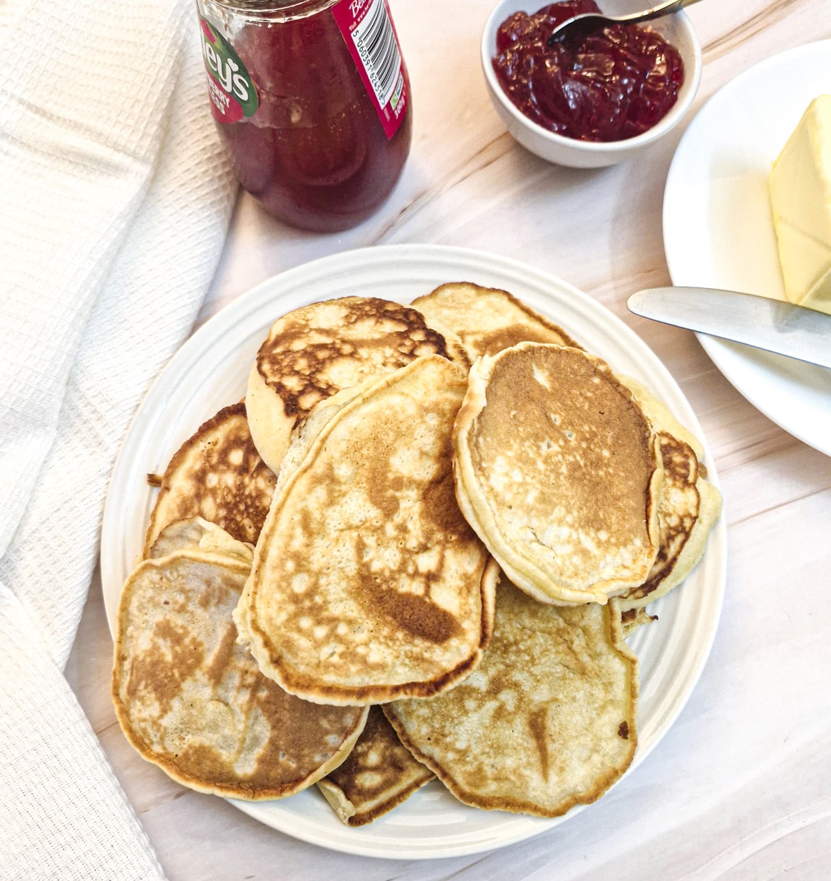 A pile of drop scones on a white plate.  There is butter and jam in the background.