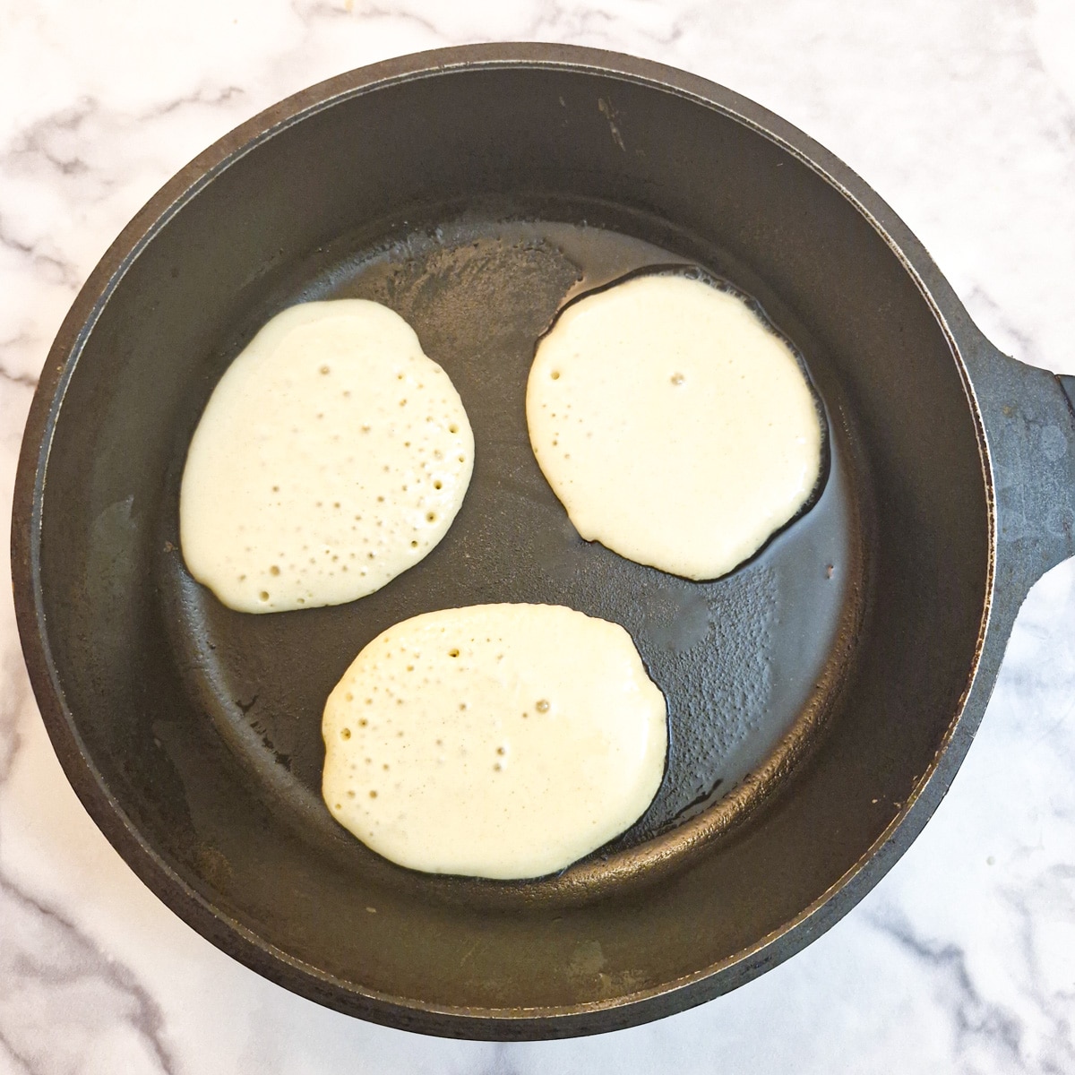 Three drop scones in a frying pan showing the bubbles forming before they are flipped.