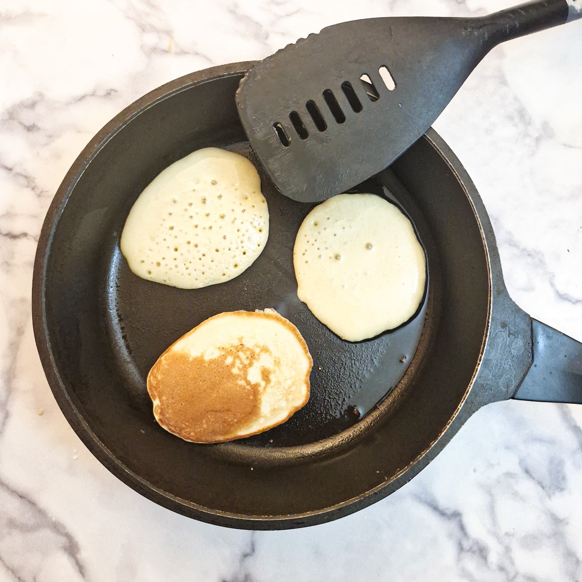 Three drop scones in a frying pan with one of them flipped over to show the browned underside.