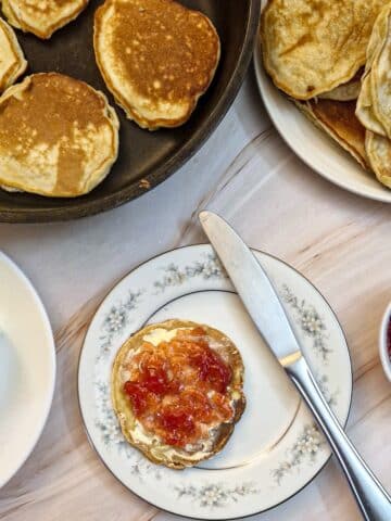 A drop scone spread with butter and jam on a white patterned plate.