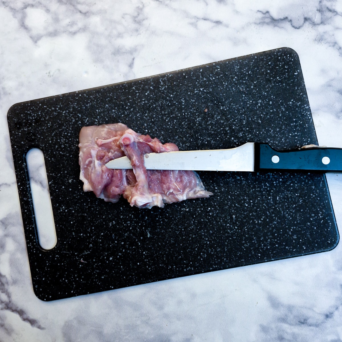 A raw chicken thigh with the meat cut away from the thigh bone and a sharp knife inserted between the bone and the meat.
