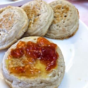 A crumpet spread with butter and strawberry jam on a plate with 3 other crumpets.