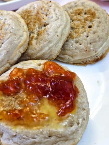 A crumpet spread with butter and strawberry jam on a plate with 3 other crumpets.