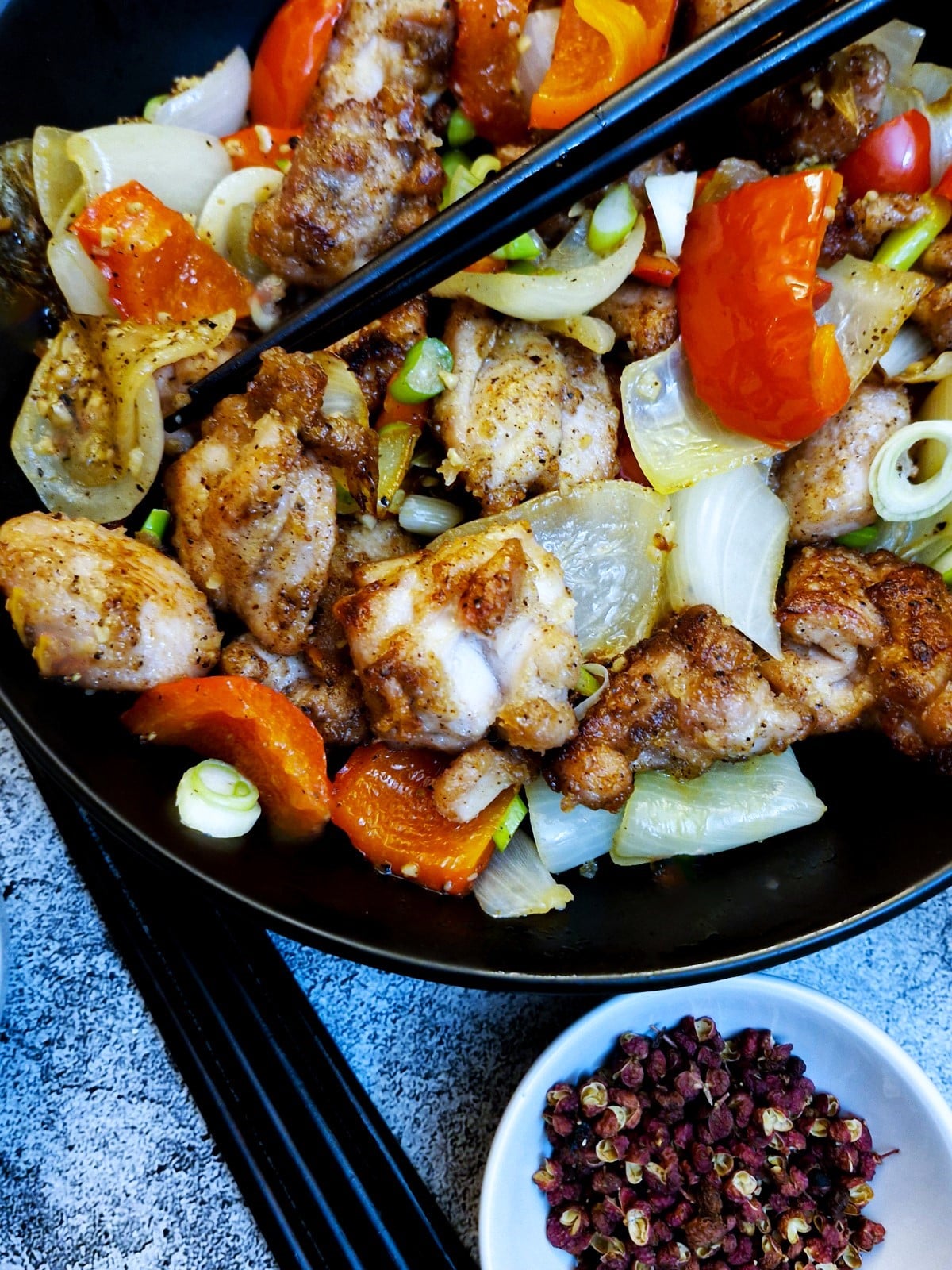 Close up of pieces of salt and pepper chicken with onions and red peppers in a black serving dish with chopsticks.  There is a bowl of Sichuan peppercons in the foreground.