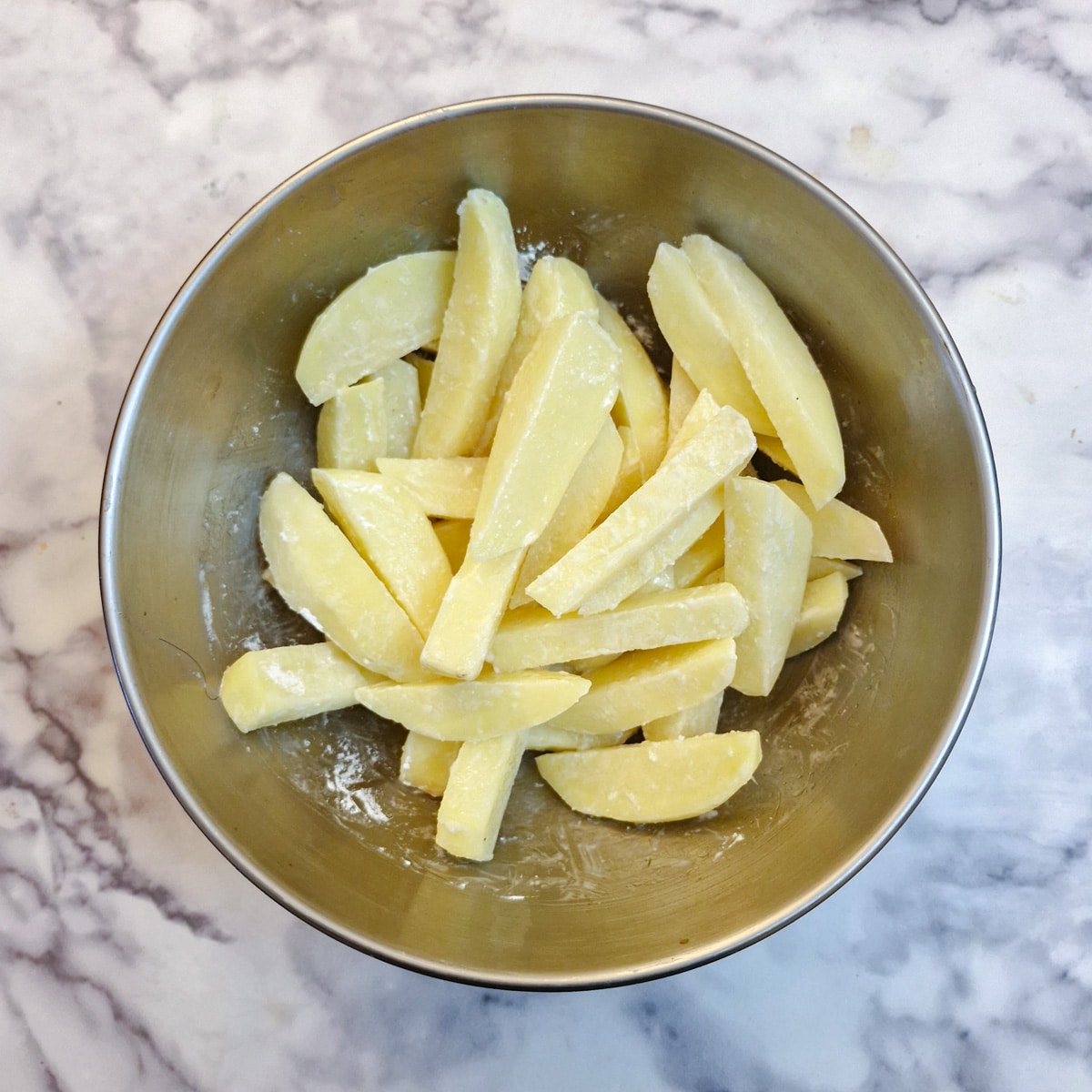 Potatoes cut into chips and coated with oil and cornflower in a metal bowl.