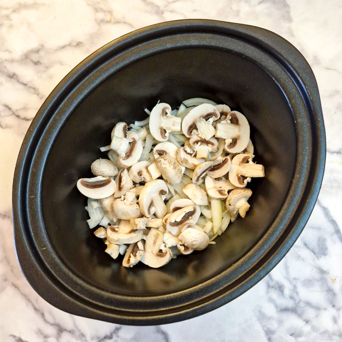 Sliced mushrooms and onions aranged on the base of a slow cooker.