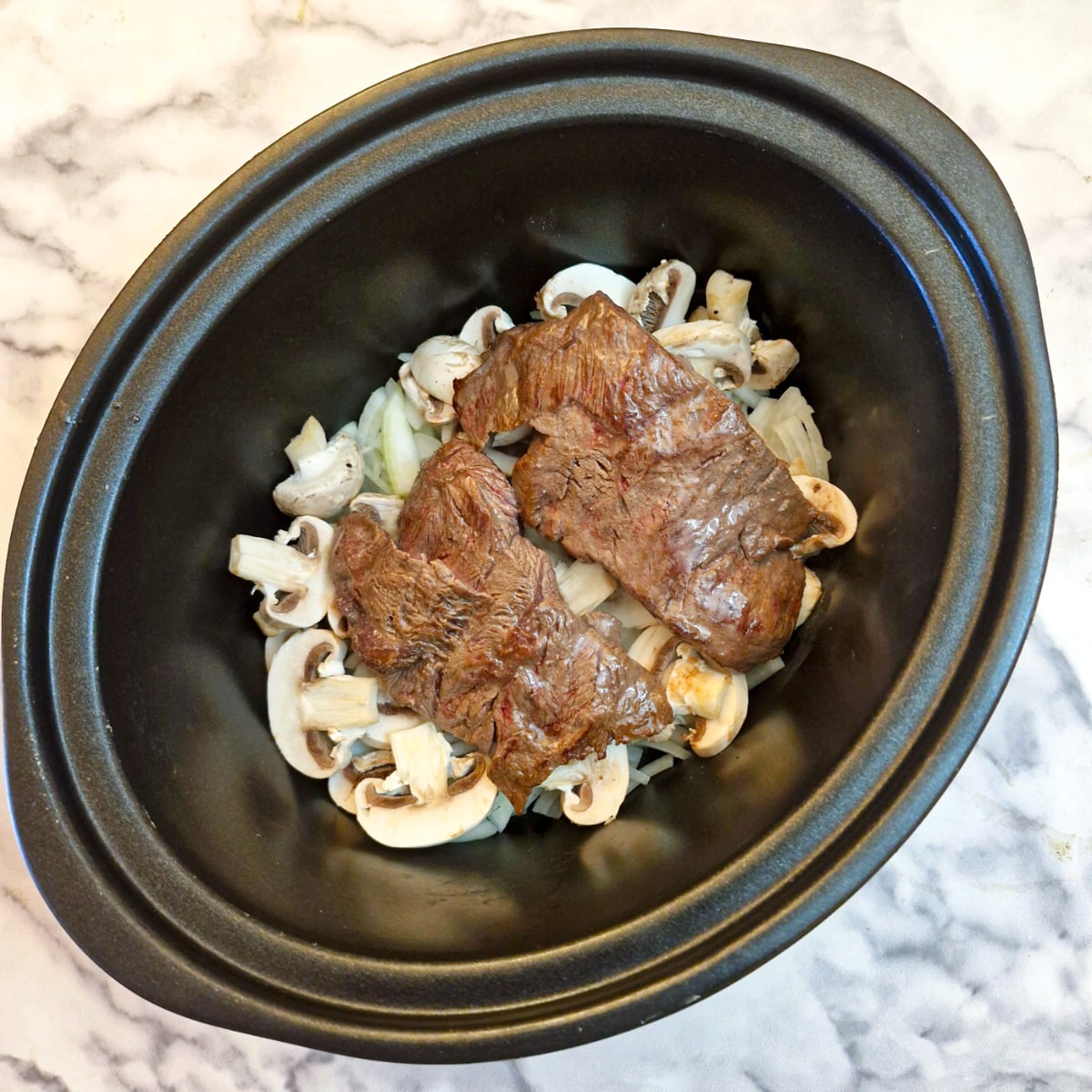 Flat iron steaks arranged on mushrooms and onions in a slow cooker.