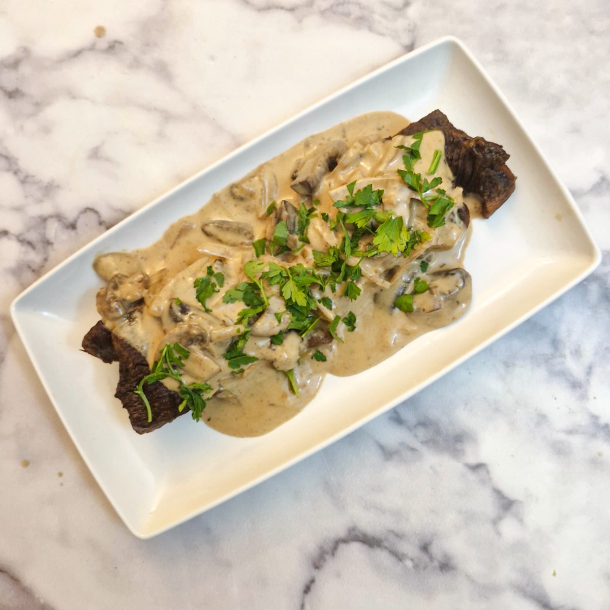 Flat iron steak covered in mushroom gravy and garnished with chopped parsley on a white oblong serving platter.