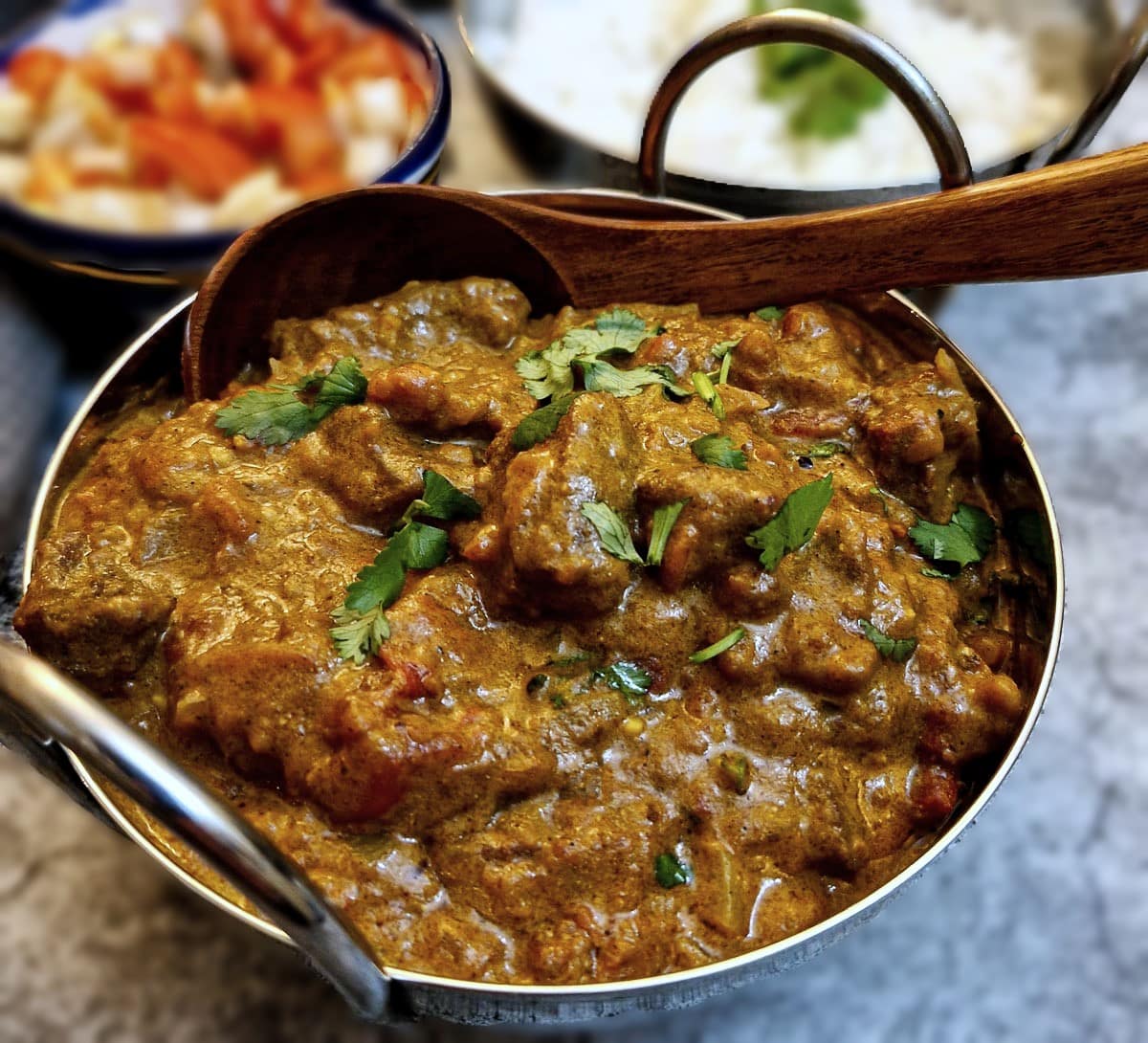 Close up of a dish of Madras beef curry.  There is a dish of rice and a dish of tomato sambal in the background.